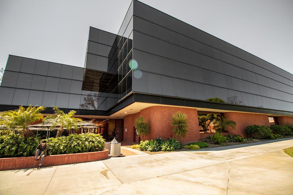 A contemporary glass building with lush plants and a person sitting outside, capturing modern architecture.